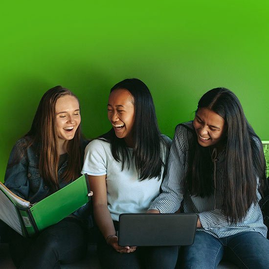 Three women sitting together, looking at a laptop against a green background
